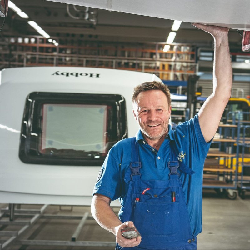 Ein Mitarbeiter von Hobby in einem blauen Overall arbeitet in der Werkshalle von Hobby, im Hintergrund sind Hobby Wohnwagen zu sehen.